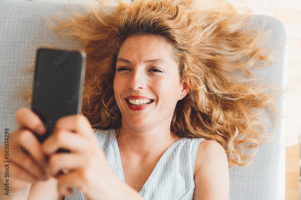 Top down view of a smiling woman laying on the couch taking on a video call, holding her phone up in the air 