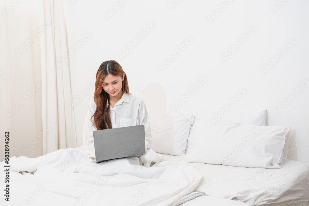 Woman in white nightgown waking up on weekend morning resting and relaxing playing with laptop mobile phone Eating bread and drinking tea in glass inside white bedroom. Morning vacation concept. Woman in white nightgown waking up on weekend morning resting and relaxing playing with laptop mobile phone Eating bread and drinking tea in glass inside white bedroom. Morning vacation concept.