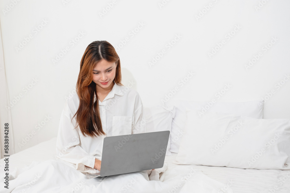 Woman in white nightgown waking up on weekend morning resting and relaxing playing with laptop mobile phone Eating bread and drinking tea in glass inside white bedroom. Morning vacation concept. Woman in white nightgown waking up on weekend morning resting and relaxing playing with laptop mobile phone Eating bread and drinking tea in glass inside white bedroom. Morning vacation concept.