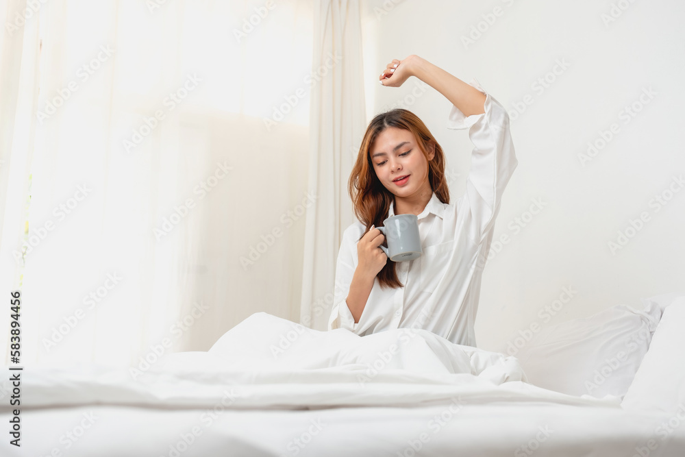 Woman in white nightgown waking up on weekend morning resting and relaxing playing with laptop mobile phone Eating bread and drinking tea in glass inside white bedroom.  Morning vacation concept.