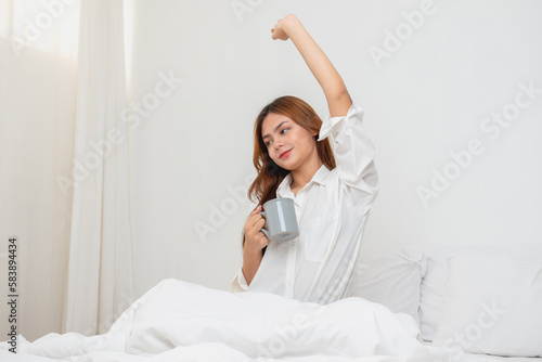 Woman in white nightgown waking up on weekend morning resting and relaxing playing with laptop mobile phone Eating bread and drinking tea in glass inside white bedroom. Morning vacation concept.
