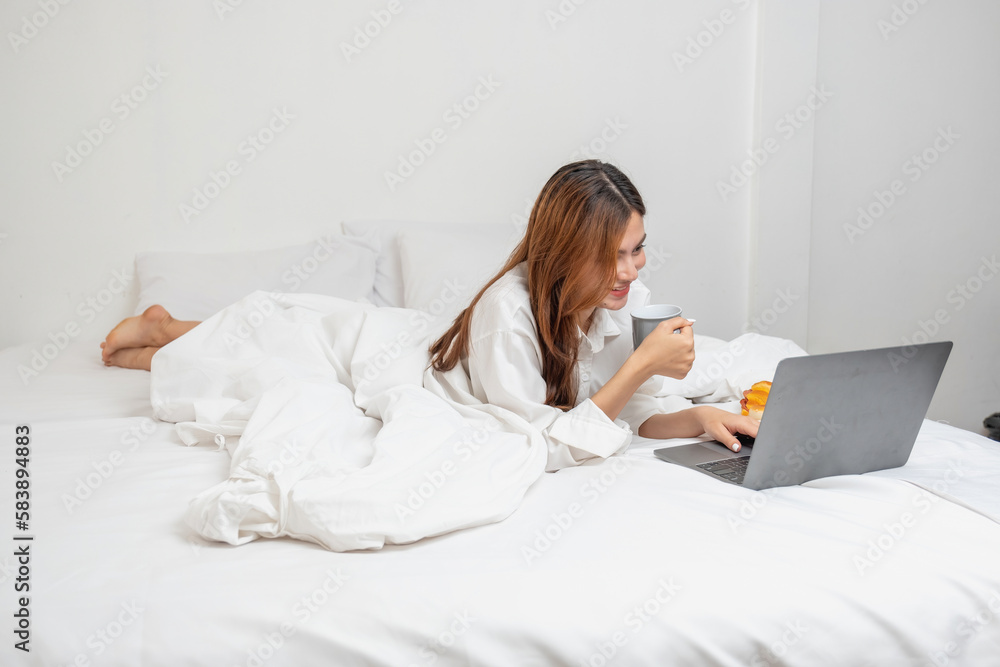 Woman in white nightgown waking up on weekend morning resting and relaxing playing with laptop mobile phone Eating bread and drinking tea in glass inside white bedroom. Morning vacation concept. Woman in white nightgown waking up on weekend morning resting and relaxing playing with laptop mobile phone Eating bread and drinking tea in glass inside white bedroom. Morning vacation concept.