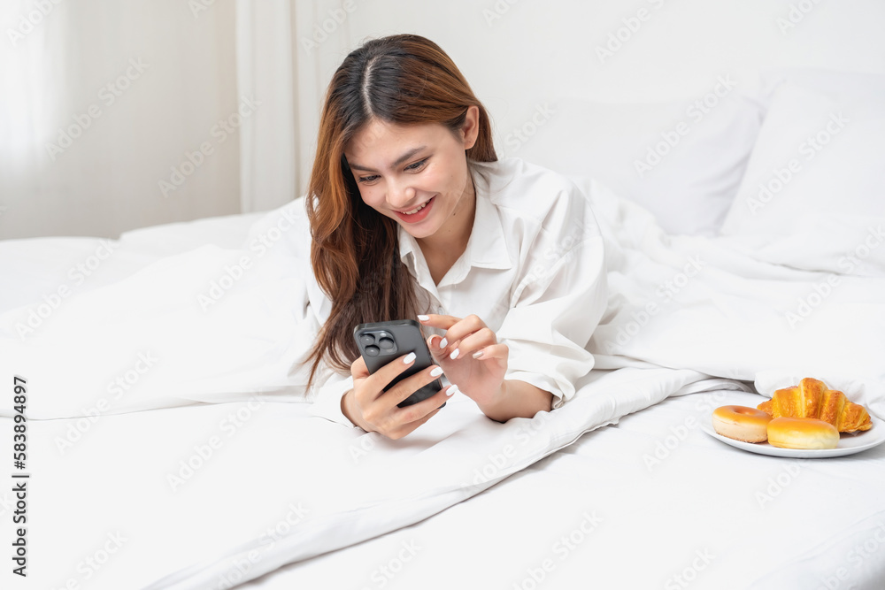 Woman in white nightgown waking up on weekend morning resting and relaxing playing with laptop mobile phone Eating bread and drinking tea in glass inside white bedroom. Morning vacation concept. Woman in white nightgown waking up on weekend morning resting and relaxing playing with laptop mobile phone Eating bread and drinking tea in glass inside white bedroom. Morning vacation concept.