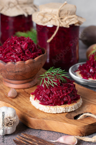 Homemade spicy caviar from beets, carrots, onions and garlic on toasted bread and also in jars on wooden board on gray background