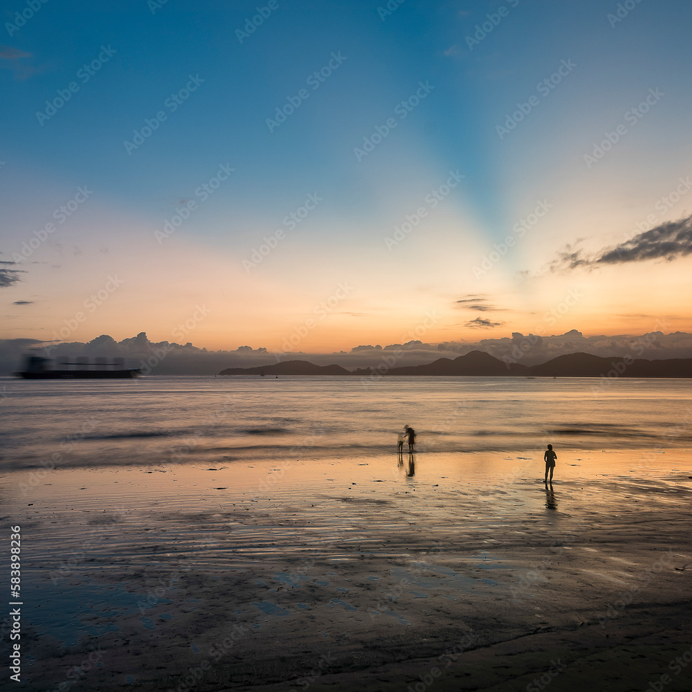 Sunset at the beach. Silhouette of people near the sea and a cargo ship passing by on the horizon. Long exposure photography. Santos, Brazil.