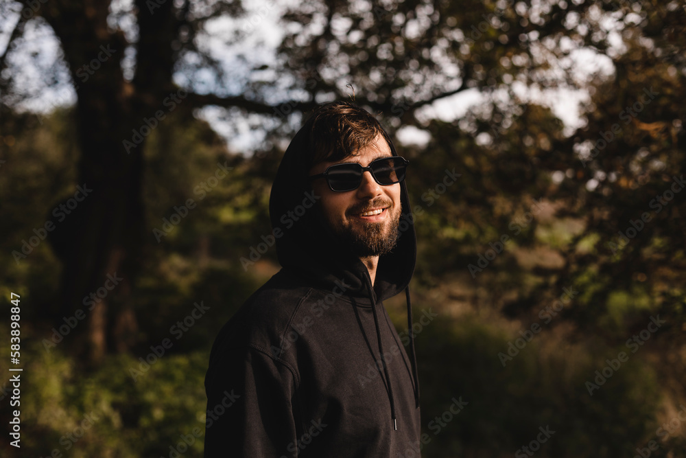 Handsome man walk in the park, he wear black glasses, hoody and hood on head. Freedom traveler concept. Happy bearded man in glasses and hood smiling and look at camera, laughing while rest in nature.