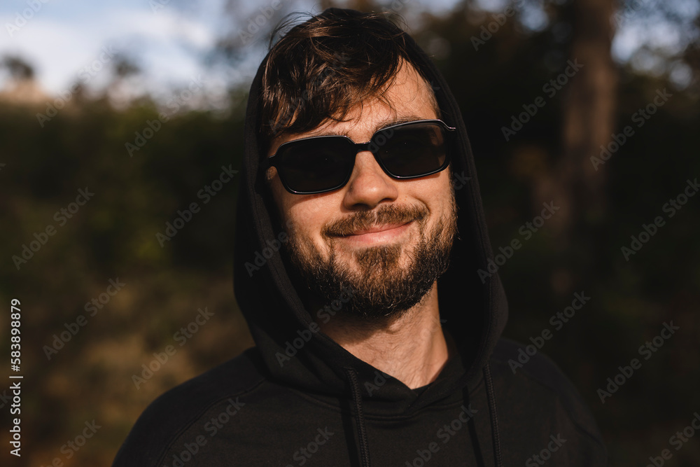 Close up handsome man walk in the park, he wear black glasses, hoody and hood on head. Freedom traveler concept. Happy bearded man in glasses and hood smiling and look at camera while rest in nature. Close up handsome man walk in the park, he wear black glasses, hoody and hood on head. Freedom traveler concept. Happy bearded man in glasses and hood smiling and look at camera while rest in nature.