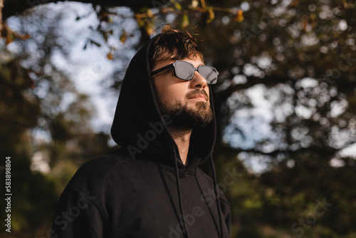 Handsome man walk in the park and look up, he wear black glasses, hoody and hood on head. Freedom traveler concept. Happy bearded man in glasses and hood smiling and looking away while rest in nature.