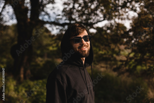 Handsome man walk in the park, he wear black glasses, hoody and hood on head. Freedom traveler concept. Happy bearded man in glasses and hood smiling and look at camera, laughing while rest in nature. © zvkate