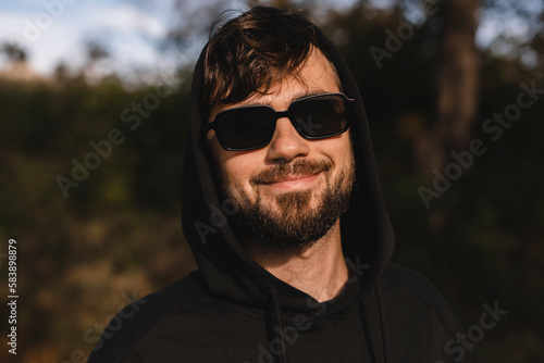 Close up handsome man walk in the park, he wear black glasses, hoody and hood on head. Freedom traveler concept. Happy bearded man in glasses and hood smiling and look at camera while rest in nature.