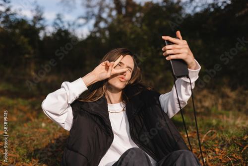 Happy young woman photographing herself using her mobile phone. Caucasian female taking selfie with her smart phone at fall park. Positive lady making photo outside show peace v sign, make blow kiss.