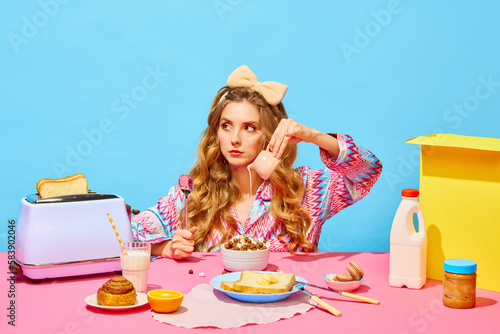 Food pop art photography. Sad young girl with long curly hair preparing milk flakes with hungry eyes over light blue background. Table with milk, butter, bread photo