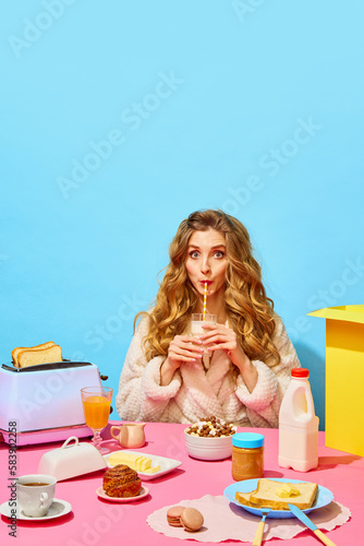 Food pop art. Charming girl wearing bathrobe drinking milkshake with pleasure facial expression over light blue background. Breakfast table setting photo