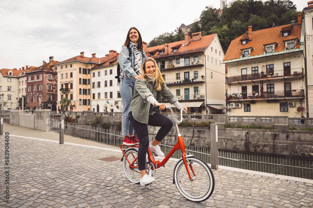 Couple of best friends riding a bike on the city streets, one of the women standing on the trunk of the bike 