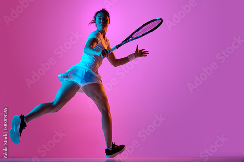 Full-length side view image of female tennis player in motion, wearing uniform, training against pink studio background in neon light. Concept of professional sport, movement, health, action. Ad