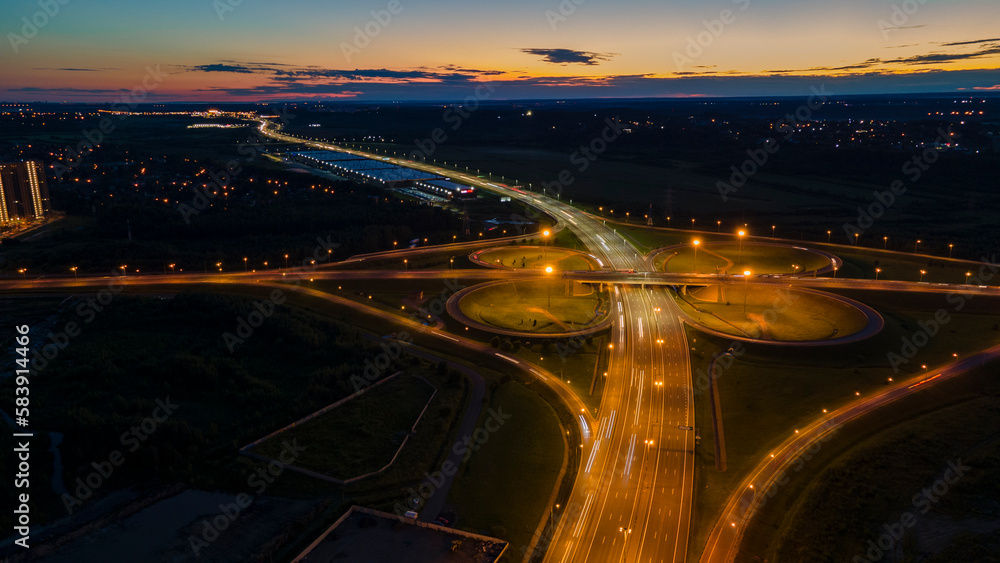 Aerial view of the bigger transportation interchange with many cars next to forest and the historical and at same time modern city of St. Petersburg at summer night