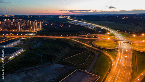 Aerial view of the bigger transportation interchange with many cars next to forest and the historical and at same time modern city of St. Petersburg at light summer night, fog © Alex