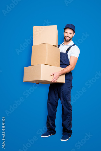 Full size fullbody portrait of attractive cheerful deliver in blue uniform with stubble, looking at camera having three big boxes in arms, isolated on grey background