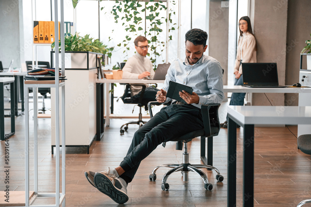 Talking behind the person that is sitting on the chair with digital device. Men and woman are working in the office together Talking behind the person that is sitting on the chair with digital device. Men and woman are working in the office together