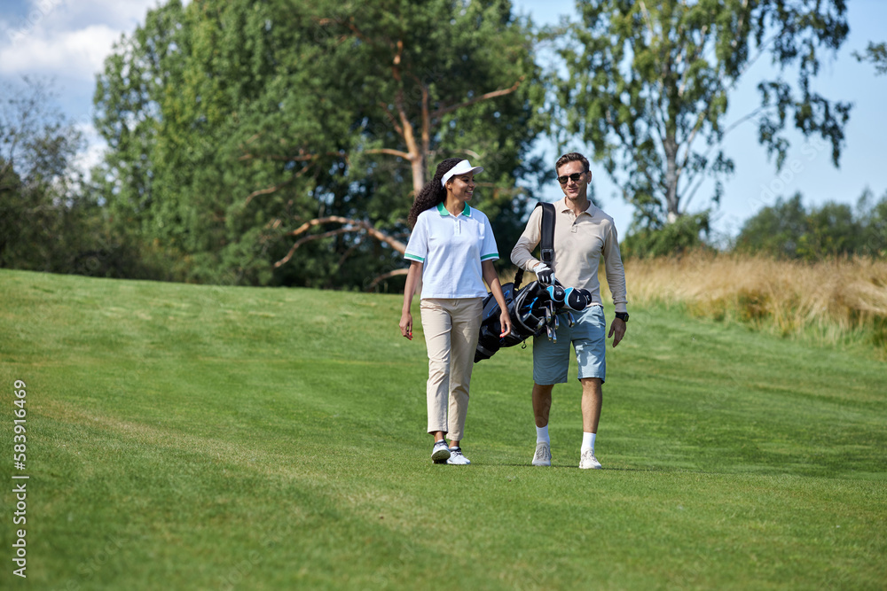 Full length portrait of elegant sporty couple carrying golf bag walking on green field towards camera and chatting, copy space Full length portrait of elegant sporty couple carrying golf bag walking on green field towards camera and chatting, copy space