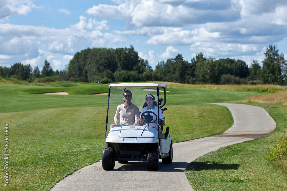 Front view of couple driving golf cart on road towards camera across golf club fields Front view of couple driving golf cart on road towards camera across golf club fields