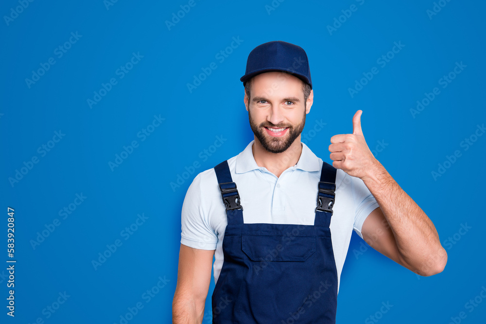 Portrait of cheerful positive deliver in blue uniform with stubble showing thumb up with finger looking at camera isolated on grey background