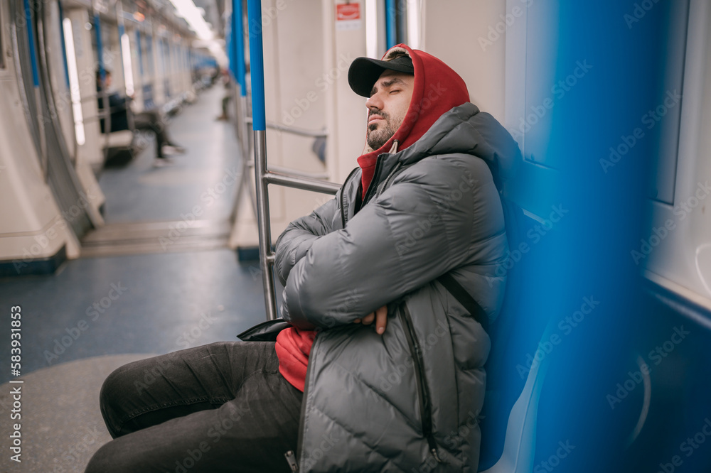 A tired man is napping in a modern subway car. A young guy in a jacket