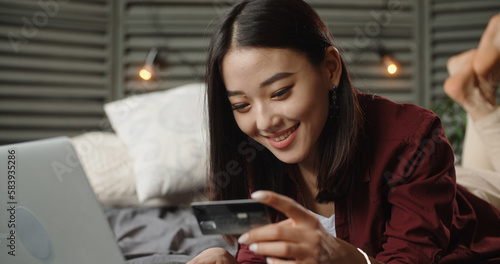 Gen Z student girl surfing online shop, attending online shopping, Young Asian Woman Spending Time on online shopping, lying on large bed with laptop, keying in credit card and positively smiling. 
