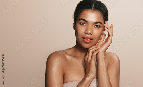 Skin care routine. Dark skinned woman with perfect glowing body, head and shoulders, touches her nourished face after skin cream and moisturizer, brown background