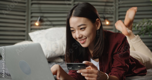 Photography Young panasian student spending time on online shopping, lying on large bed with