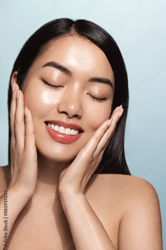 Vertical shot of smiling korean girl washes her face, applied skincare routine, facial cream with pleased face, touches her soft skin, stands topless against blue background Vertical shot of smiling korean girl washes her face, applied skincare routine, facial cream with pleased face, touches her soft skin, stands topless against blue background
