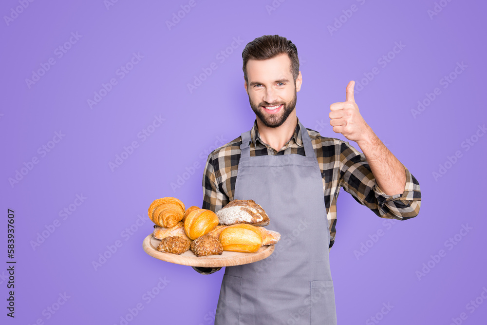 Portrait of positive attractive baker in uniform with stubble holding tray with bakery products in hand, showing thumb up with finger, isolated on grey background Portrait of positive attractive baker in uniform with stubble holding tray with bakery products in hand, showing thumb up with finger, isolated on grey background