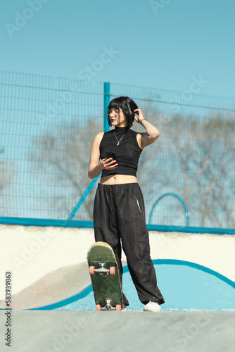 Young pretty skater girl posing with her skateboard and using her phone in the skate park
