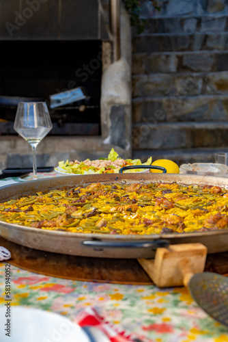 Paellera with a freshly cooked authentic Valencian paella, to serve on a table with the dishes set, salad, glasses and the tablecloth with floral motifs with a rustic barbecue in the background.