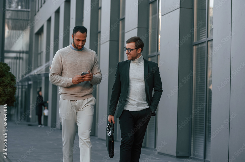Holding smartphone. Businessman in formal clothes is with black man outdoors in the city Holding smartphone. Businessman in formal clothes is with black man outdoors in the city