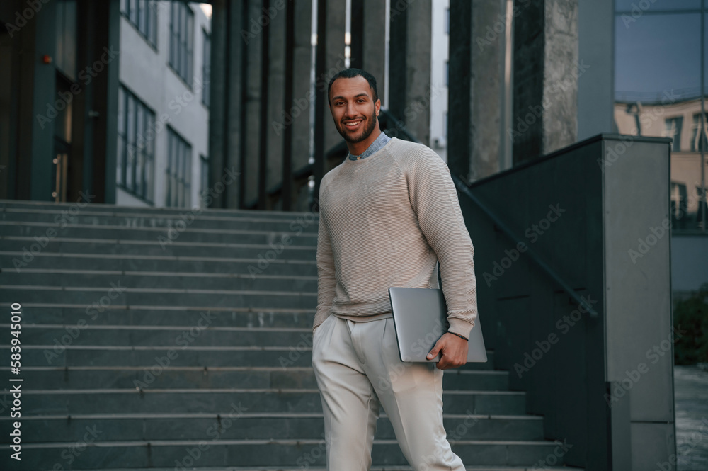 Fototapeta premium With laptop in hands. Handsome black man is outdoors near the business building