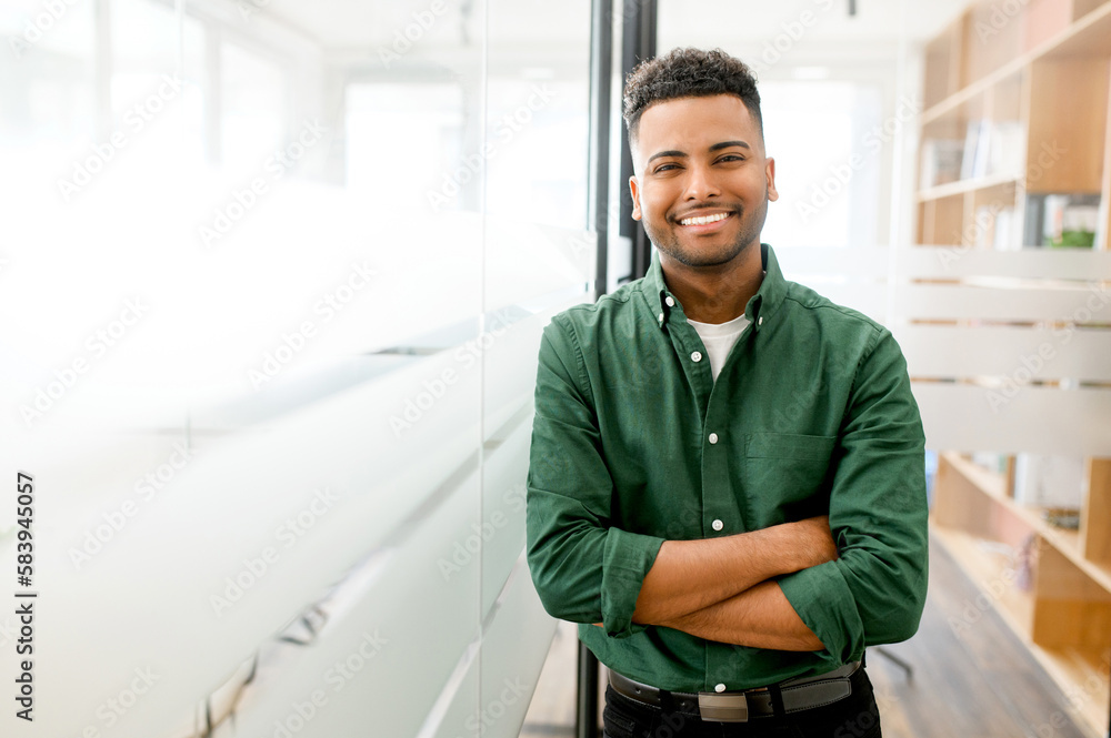 Handsome indian male office employee standing with his arms crossed, looking at the camera and laughs. Portrait of cheerful young man wearing smart casual shirt in modern coworking space - obrazy, fototapety, plakaty 