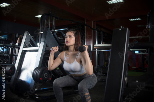 A fit young asian woman does dumbbell squats, holding the dumbbells in the front rack position. Training legs at a modern gym.