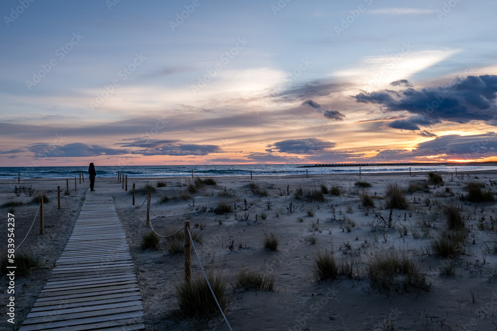 Persona de lejos en una pasarela de madera contemplando una bonita puesta de sol en una desertica playa.
