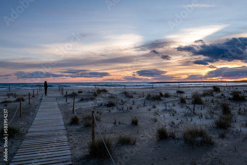 Persona de lejos en una pasarela de madera contemplando una bonita puesta de sol en una desertica playa.