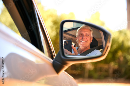 Senior Male Passenger With Mobile Phone Reflected In Wing Mirror Of Car Enjoying Day Trip Out