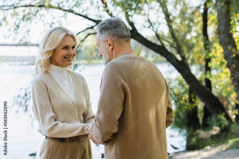 happy middle aged woman holding hands with husband while looking at each other near lake in park. happy middle aged woman holding hands with husband while looking at each other near lake in park.