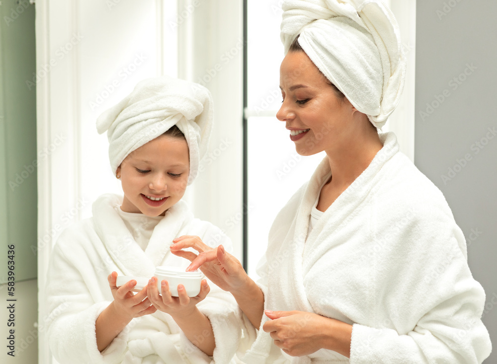 Smiling caucasian small daughter and little young mom in bathrobe and towel, have fun, apply cream Smiling caucasian small daughter and little young mom in bathrobe and towel, have fun, apply cream