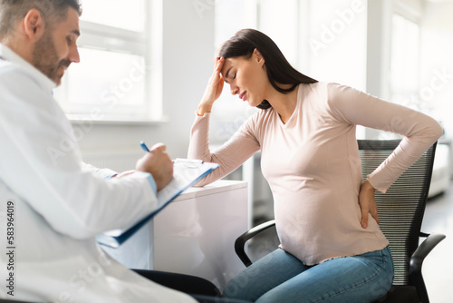 Sick pregnant lady feeling unwell  touching head and back during meeting with her doctor in clinic