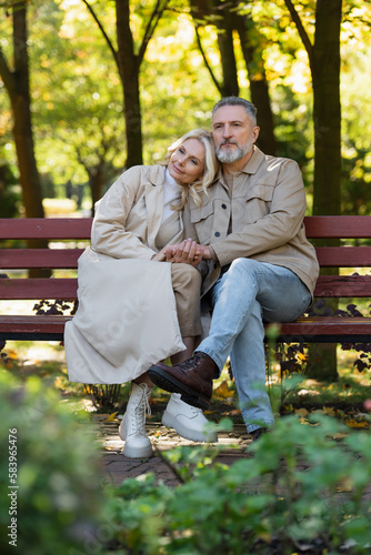 Blonde woman holding hand of mature husband while sitting on bench in spring park.