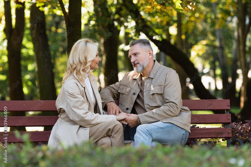 Mature man talking and holding hand of blonde wife while sitting on bench in park.