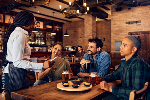 Group of young people choosing order from digital menu while talking to waitress in pub.