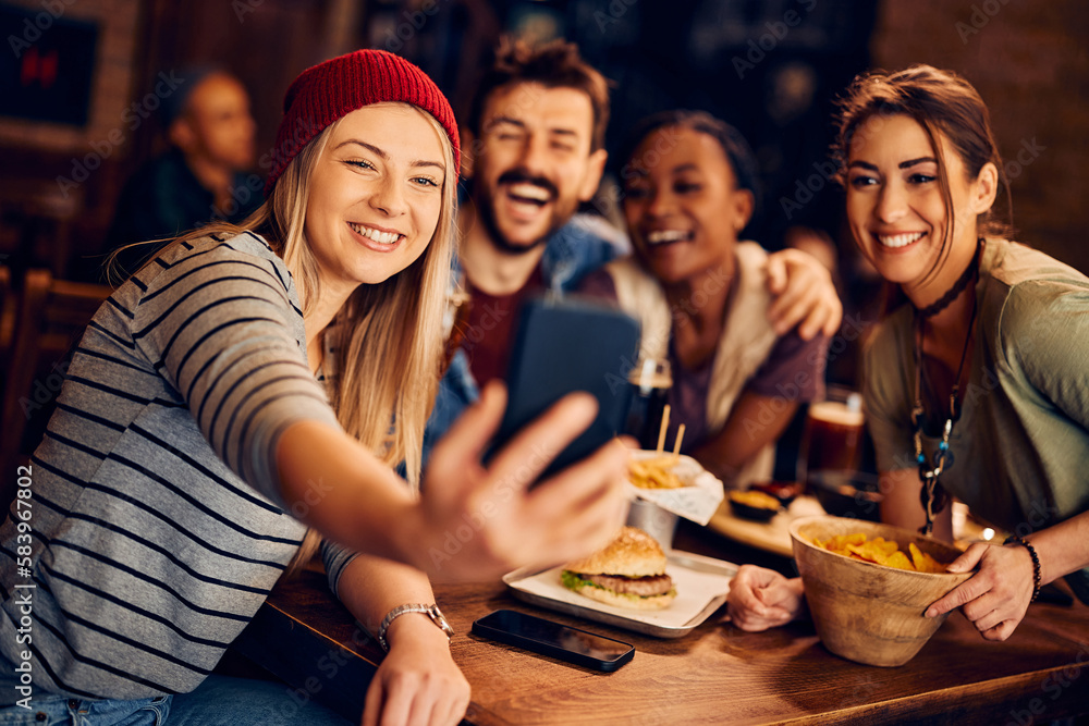 Happy woman using smart phone while taking selfie with friends in pub.