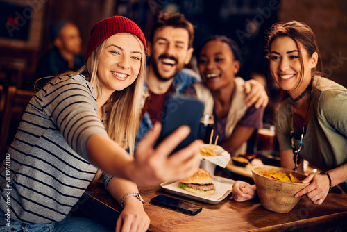 Happy woman using smart phone while taking selfie with friends in pub.