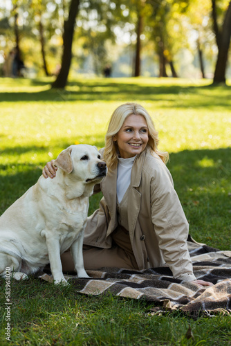 happy middle aged woman with blonde hair cuddling labrador dog while sitting on blanket in park.